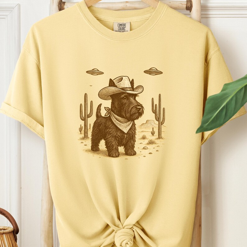 May include: Yellow t-shirt featuring a cartoon illustration of a Scottish Terrier wearing a cowboy hat and bandana, set against a desert landscape with cacti and UFOs. The shirt has a Comfort Colors tag.