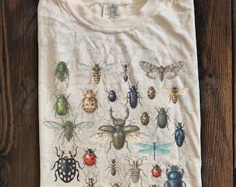 May include: An ivory-colored t-shirt featuring a detailed graphic print of various insects, including beetles, a dragonfly, a moth, and ants. The shirt has a crew neckline and a Comfort Colors brand label printed on the inside of the collar.