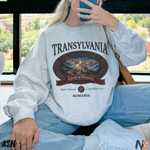 Puede incluir: Una sudadera gris claro de cuello redondo con un gr&aacute;fico ovalado marr&oacute;n oscuro que muestra una ilustraci&oacute;n detallada de un castillo. El texto dice TRANSYLVANIA, Castle of Dracula, Vlad's Domain, Carpathian Lore y ROMANIA en una fuente serif marr&oacute;n oscuro.