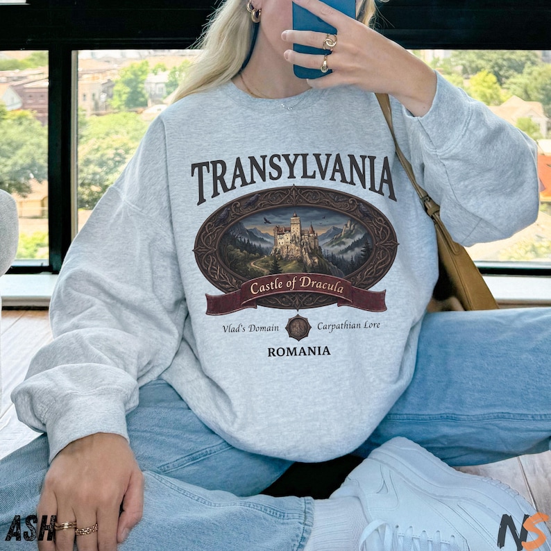 Puede incluir: Una sudadera gris claro de cuello redondo con un gr&aacute;fico ovalado marr&oacute;n oscuro que muestra una ilustraci&oacute;n detallada de un castillo. El texto dice TRANSYLVANIA, Castle of Dracula, Vlad's Domain, Carpathian Lore y ROMANIA en una fuente serif marr&oacute;n oscuro.
