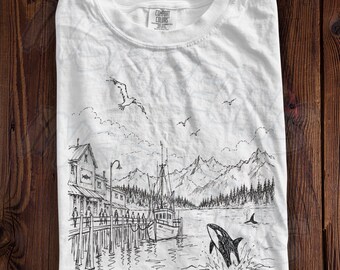 May include: A white crew neck t-shirt featuring a black ink-style illustration of a coastal harbor scene. The graphic depicts a wooden pier with buildings, a fishing boat, mountains in the background, birds in the sky, and an orca whale breaching.