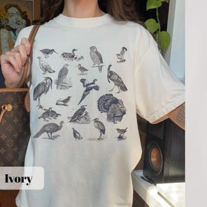 May include: Ivory-colored t-shirt featuring a collection of detailed bird illustrations in shades of gray. The shirt has a relaxed fit and short sleeves. The word "Ivory" is displayed in a white rectangle at the bottom of the image.
