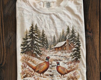 May include: An ivory crew neck t-shirt featuring a graphic print of two pheasants standing in a snowy landscape with a log cabin and pine trees in the background. The inner neck label displays the Comfort Colors brand name and text.