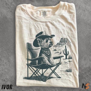 May include: Ivory-colored t-shirt featuring a graphic of a dog wearing a cowboy hat and bandana, seated in a folding chair. The design includes a desert landscape with a cactus and rock formations. The shirt is labeled "IVOR" and "NS".