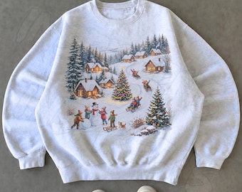 May include: A light gray crewneck sweatshirt featuring a printed winter scene with snow-covered cabins, pine trees, a decorated Christmas tree, and children playing in the snow with a snowman, sleds, and a small dog in the foreground.