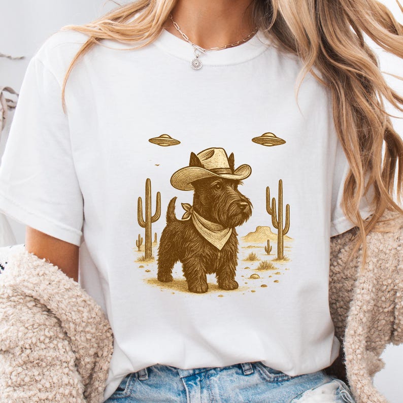 May include: White t-shirt featuring a brown illustration of a Scottish Terrier wearing a cowboy hat and bandana, set against a desert landscape with cacti and UFOs. The design has a vintage, Western theme.