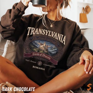 Puede incluir: Una sudadera de cuello redondo en color marr&oacute;n chocolate oscuro con un estampado gr&aacute;fico que incluye el texto TRANSYLVANIA, Castle of Dracula, Vlad's Domain, Carpathian Lore y ROMANIA. El dise&ntilde;o presenta un marco ovalado con un castillo.