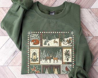 May include: An olive green crewneck sweatshirt featuring a rectangular graphic print composed of multiple panels. The design displays woodland scenes with a fox, a rabbit, a squirrel, deer, pine trees, a lantern, pinecones, and a cardinal.