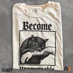 May include: Ivory t-shirt featuring a black graphic of a cat biting an arm, within a rectangle. The word "Become" is above the graphic, and "Unmaamormable" is below. The shirt has a Comfort Colors tag.