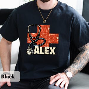May include: Black t-shirt with a red cross design, a stethoscope graphic, and the name "ALEX" in white. The shirt has a crew neck and is made of a soft material.