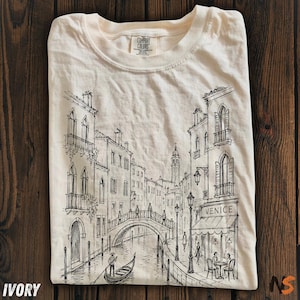 May include: An ivory short-sleeve t-shirt featuring a black ink-style line drawing of a Venetian canal scene. The graphic depicts buildings, a bridge, a gondola with a gondolier, and a cafe with the text "VENICE" printed on the awning.
