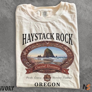 Peut inclure: T-shirt ivoire avec un motif de Haystack Rock, un point de repère côtier de l'Oregon. Le motif comprend un cadre ovale avec des coquillages, des oiseaux et les mots "Coastal Discovery". Le t-shirt porte également les mots "Oregon" et "Pacific Legacy".