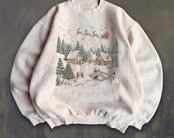 May include: A cream-colored crewneck sweatshirt featuring a printed winter scene with houses, pine trees, a stone bridge, a snowman, and Santa Claus in a sleigh pulled by reindeer flying across a starry night sky with a crescent moon.