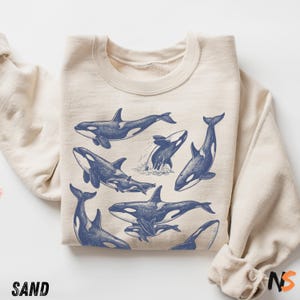 May include: A sand-coloured sweatshirt with a navy blue illustration of orcas. The orcas are shown in different positions, including breaching the water. The word "SAND" is printed in the lower left corner, and "NS" is printed in the lower right corner.