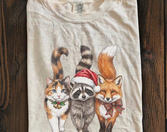 May include: An ivory t-shirt featuring a graphic of a calico cat with a bell collar, a raccoon wearing a red and white Santa hat, and a red fox wearing a green and red plaid bow. The inner neck label reads Comfort Colors.