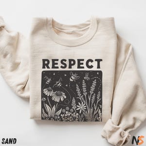 May include: A sand-colored crewneck sweatshirt featuring the word RESPECT in bold black capital letters above a black rectangular graphic. The illustration depicts white line-art bees flying among various wildflowers, including daisies and lavender stalks.
