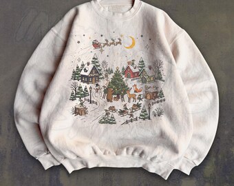 May include: A cream-colored crewneck sweatshirt featuring a detailed illustration of a winter village scene with houses, pine trees, a decorated Christmas tree, a bear, a deer, and Santa Claus flying in a sleigh across a crescent moon.
