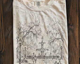 May include: An ivory-colored t-shirt featuring a black ink-style illustration of a park scene with a central fountain, large trees with hanging moss, street lamps, and several small figures of people walking or sitting on benches.