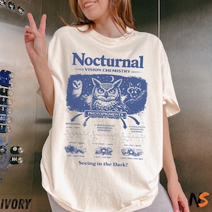 Może przedstawiać: Kremowa koszulka z niebieską grafiką przedstawiającą sowę, szopa pracza oraz tekst "Nocturnal Vision Chemistry" i "Seeing in the Dark!". Projekt zawiera schematy naukowe.