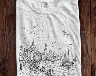 May include: A white short-sleeve t-shirt featuring a black ink sketch of the Boston, Massachusetts waterfront. The graphic depicts a harbor with sailboats, a city skyline, and people walking along the shore. The text reads Boston, Massachusetts.