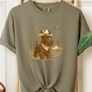 May include: Olive green t-shirt featuring a cartoon illustration of a black dog wearing a cowboy hat and bandana, with two UFOs above. The dog stands in a desert scene with cacti. The shirt has the Comfort Colors logo.