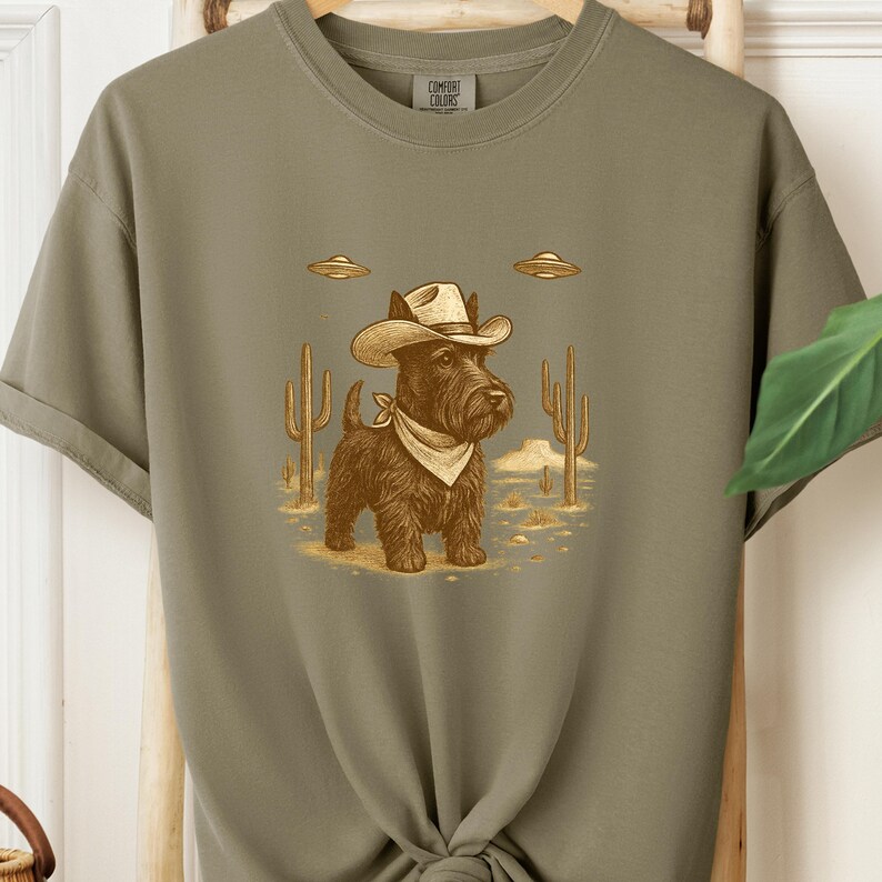 May include: Olive green t-shirt featuring a cartoon illustration of a black dog wearing a cowboy hat and bandana, with two UFOs above. The dog stands in a desert scene with cacti. The shirt has the Comfort Colors logo.