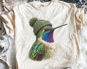 May include: An ivory short-sleeve t-shirt featuring a graphic of a hummingbird wearing a green knit beanie with a pom-pom. The bird has iridescent green, blue, and purple feathers on its head and neck with a white chest and long beak.