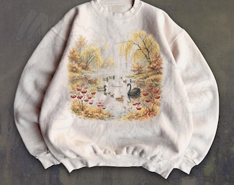 May include: A light beige crewneck sweatshirt featuring a central graphic print of a pond scene with a black swan, several ducks, and willow trees surrounded by autumn foliage with red berries in a soft, painterly artistic style.