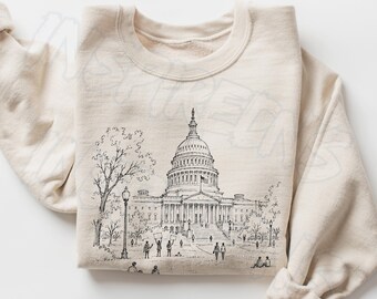 May include: A sand-colored crewneck sweatshirt featuring a black ink-style line drawing of the United States Capitol building. The illustration depicts the dome, columns, surrounding trees, and small figures of people on the grounds.