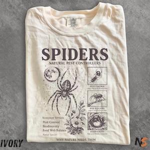 May include: Ivory-colored t-shirt with a graphic design featuring a spider, moon, and botanical elements. Text includes "SPIDERS NATURAL PEST CONTROLLERS" and "WHY NATURE NEEDS THEM". The shirt also lists ecosystem services.
