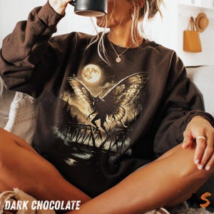 May include: A dark chocolate brown crewneck sweatshirt featuring a graphic print of a winged creature standing on a bridge under a full moon. The design includes silhouettes of trees and a starry night sky, rendered in shades of gold and black.