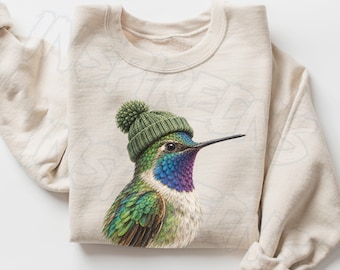 May include: A sand-colored crewneck sweatshirt featuring a graphic of a hummingbird wearing a green knit beanie with a pom-pom. The bird has iridescent green, blue, and purple feathers on its head and chest, with a long, thin black beak.