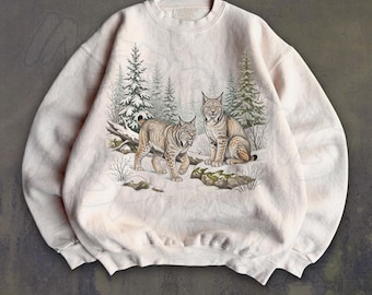 May include: A light beige crewneck sweatshirt featuring a graphic print of two lynx standing in a snowy forest landscape with evergreen trees. The illustration depicts one lynx walking and another sitting on a rocky, snow-covered ground.