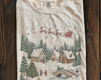 May include: An ivory t-shirt featuring a graphic print of a winter village scene with snow-covered houses, a decorated Christmas tree, a stone bridge, a snowman, and Santa Claus in a sleigh pulled by reindeer flying across a starry sky.