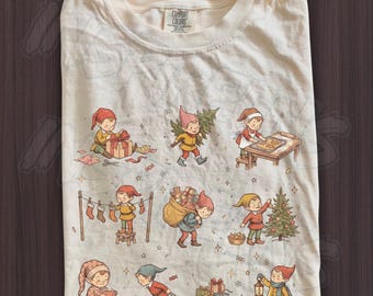 May include: A cream-colored t-shirt featuring a graphic print of various elves engaged in holiday activities, including wrapping gifts, baking cookies, hanging stockings, decorating a Christmas tree, and pulling a sled with presents.