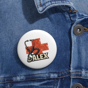 May include: A white badge featuring a red cross and a black stethoscope illustration. The name "ALEX" is printed in bold black letters below the stethoscope. The badge is round and attached to a denim jacket.