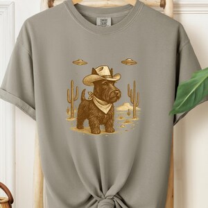 May include: A gray Comfort Colors t-shirt featuring a cartoon illustration of a black dog wearing a cowboy hat and bandana, set against a desert landscape with cacti and UFOs.