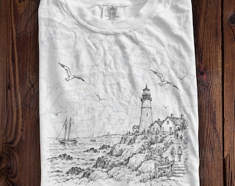 May include: A white short-sleeve t-shirt featuring a black ink-style illustration of a lighthouse on a rocky coast, a sailboat, and flying seagulls. The text Portland, Maine is printed at the bottom. The shirt has a Comfort Colors label.