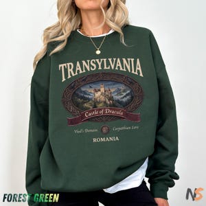 Puede incluir: Una sudadera de manga larga color verde bosque con un estampado gr&aacute;fico de un castillo dentro de un marco ovalado. El texto dice TRANSYLVANIA, Castle of Dracula, Vlad's Domain, Carpathian Lore y ROMANIA en fuente color crema al frente.