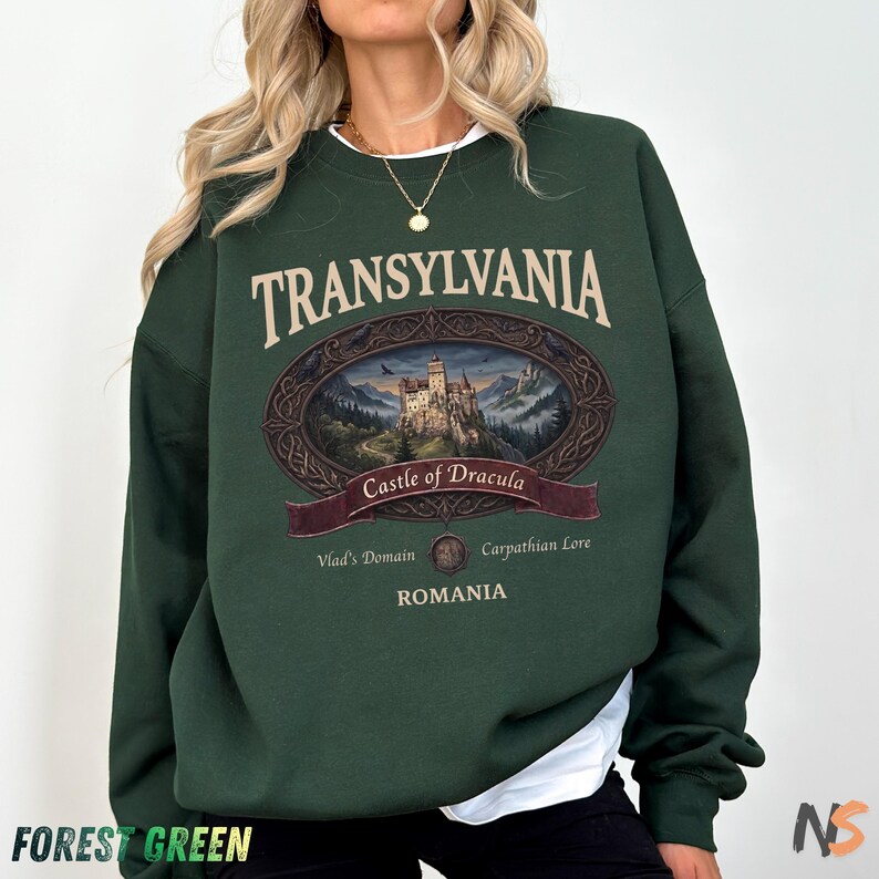 Puede incluir: Una sudadera de manga larga color verde bosque con un estampado gr&aacute;fico de un castillo dentro de un marco ovalado. El texto dice TRANSYLVANIA, Castle of Dracula, Vlad's Domain, Carpathian Lore y ROMANIA en fuente color crema al frente.