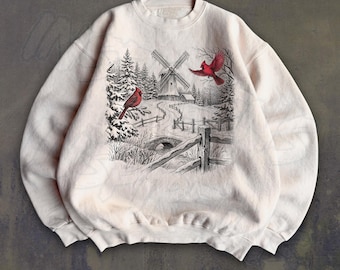 May include: A cream-colored crewneck sweatshirt featuring a graphic print of a winter landscape. The scene includes a windmill, snow-covered pine trees, a wooden fence, and two bright red cardinal birds perched in the snowy setting.