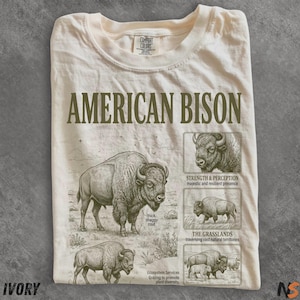 May include: An ivory-colored t-shirt featuring the text AMERICAN BISON in large green letters. The shirt displays detailed illustrations of bison in various poses, accompanied by descriptive text labels including STRENGTH & PERCEPTION, THE GRASSLANDS, and thick shaggy coat.
