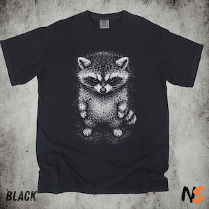 May include: A black short-sleeve t-shirt featuring a detailed white graphic illustration of a standing raccoon centered on the chest. The raccoon is depicted with textured fur, pointed ears, and a striped tail, rendered in a sketch-like style.