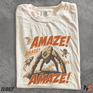 May include: Ivory-colored t-shirt with a retro space-themed graphic. The design features the word "AMAZE!" in orange, a rocket, a robot, an astronaut, and a moon. The shirt is made of Comfort Colors material.