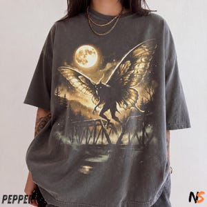 May include: A charcoal gray short-sleeve t-shirt featuring a graphic print of a winged creature flying over a bridge at night under a full moon. The design includes silhouettes of pine trees and a misty landscape in shades of gold and black.