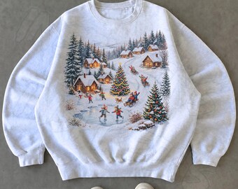 May include: A light gray crewneck sweatshirt featuring a detailed winter scene print. The graphic depicts a snowy village with log cabins, pine trees, a decorated Christmas tree, and figures ice skating and sledding on a frozen pond.