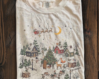 May include: A cream-colored t-shirt featuring a festive winter scene with a Christmas tree, houses, a snowman, and woodland animals. Santa Claus and his reindeer fly across the sky near a crescent moon. The shirt has a Comfort Colors brand label.