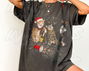 May include: A charcoal gray oversized t-shirt featuring a graphic of three cats celebrating. One cat wears a Santa hat, another has tinsel, and a third is partially visible. A champagne bottle and a red plastic cup are on the floor.