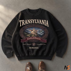 Puede incluir: Una sudadera negra de cuello redondo con un estampado gr&aacute;fico que incluye el texto TRANSYLVANIA en la parte superior, un marco ovalado con una ilustraci&oacute;n de un castillo, y el texto Castle of Dracula, Vlad's Domain, Carpathian Lore y ROMANIA.