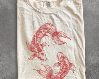 May include: Ivory-colored t-shirt featuring a red illustration of two koi fish. The shirt has a crew neck and short sleeves. The text "IVORY" is at the bottom left corner. The brand label "COMFORT COLORS" is visible at the top.